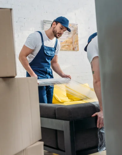 Workers preparing furniture for intercity relocation in North Shore by wrapping a sofa with protective film in a home setting surrounded by moving boxes.